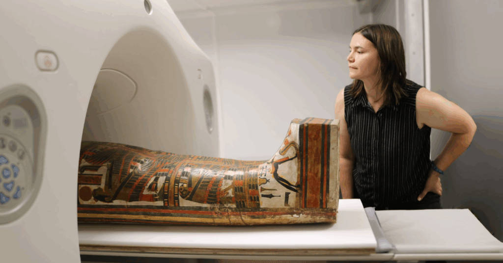 A close up horizontal shot shows a mummified body in a painted wooden coffin. The coffin is being moved into the opening of a large, modern CT scanner. A person in a striped top and black pants stands to the right, looking on with their hand on their hip. The mummy is identified as Chenet aa, and the person is a Field Museum staff member. The image captures the intersection of ancient history and modern medical technology.