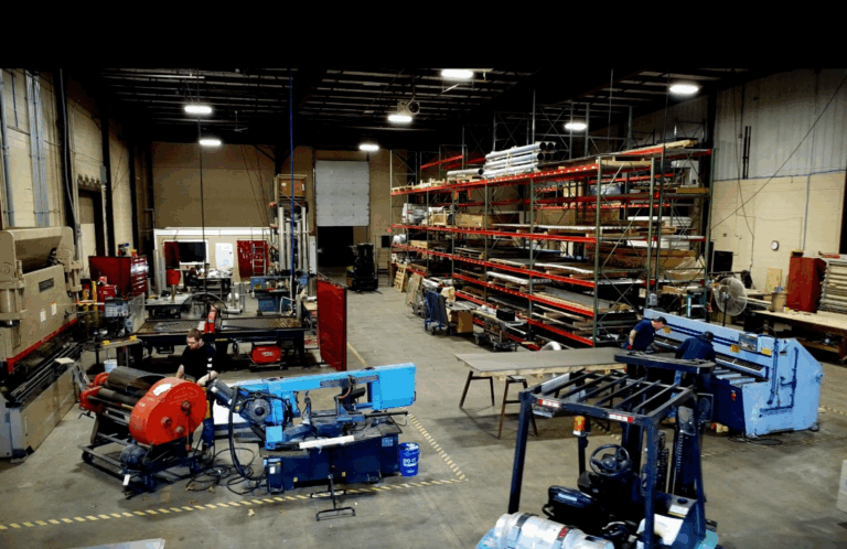 Wide shot of an industrial fabrication shop floor with various metalworking equipment, sheet metal storage racks, and workers, highlighting the facility's capability for custom MRI and RF shielding fabrication.