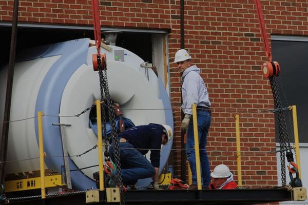 Workers securing heavy GE MRI machine for crane lift into hospital or clinic through large wall opening.