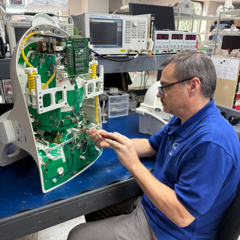 A technician wearing glasses and a blue MXR polo shirt repairs the exposed green circuit board inside a piece of a MRI Coil.