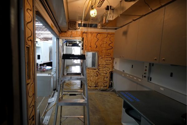 Interior view of a mobile medical trailer under construction, showing bare walls, a ladder, and overhead cabinets.
