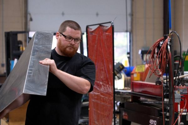 MXR fabrication worker in glasses and beard carrying large, folded sheet of bright aluminum or galvanized metal in shop.