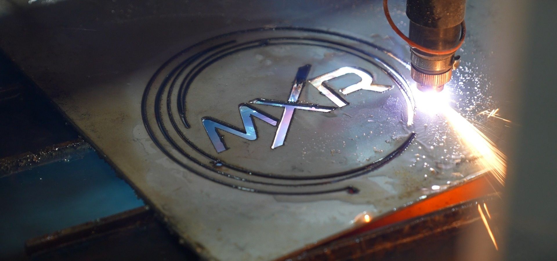 Close up of a high power plasma cutter tool etching or cutting the MXR company logo into a thick sheet of metal in a workshop.